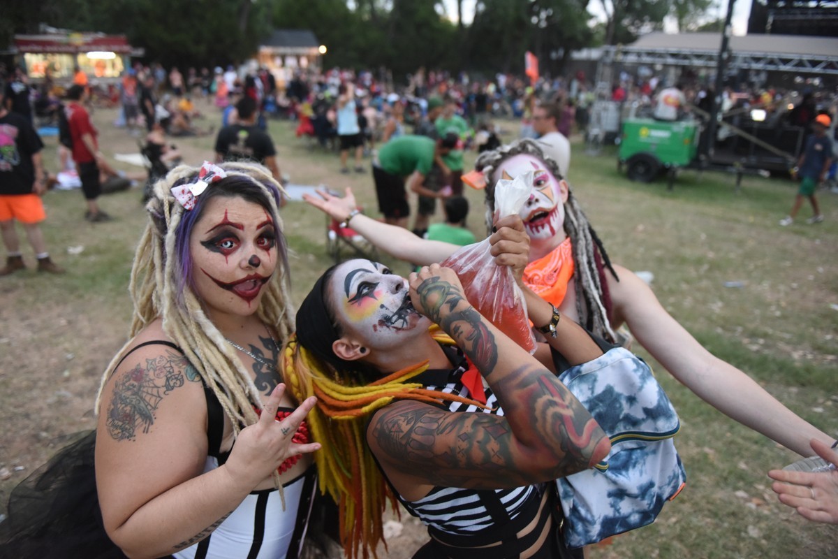 a group of three juggalos at a festival. the left woman has her hand up with three fingers showing. the middle woman has tattoes and is miming something. the right woman is partially obscured and has her arms out as if to say tada.jpg