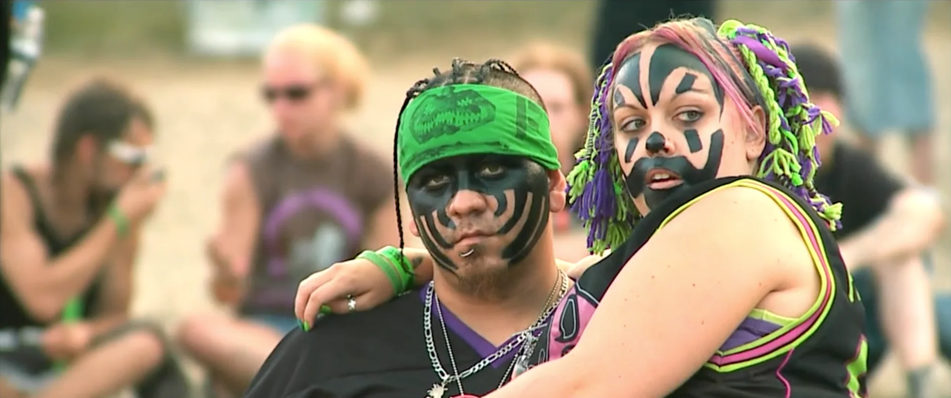 a juggalo wearing a green bandana on his head holds his juggalo wife who is wearing colourful hair extensions and a black jersey.webp