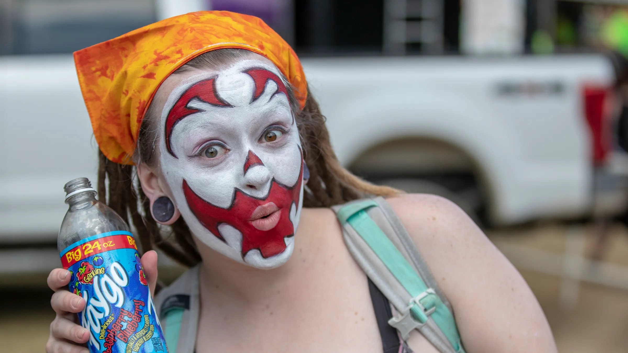 a juggalo woman enthusiastically holds a purple faygo beverage. she is wearing a bright orange bandana and red face makeup. her shoulders are exposed and there is a portion of a pick-up truck bed visible in the blurry background,.webp